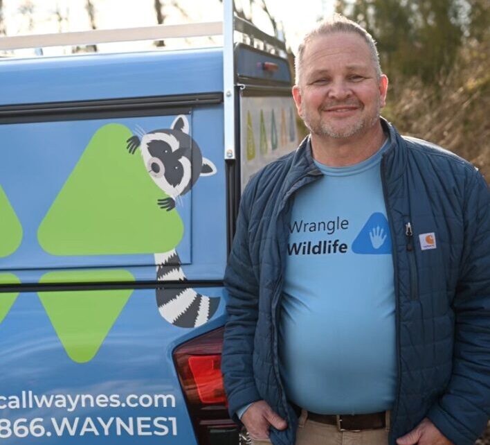 Smiling man standing beside a wildlife removal van with a cartoon raccoon graphic and visible company URL